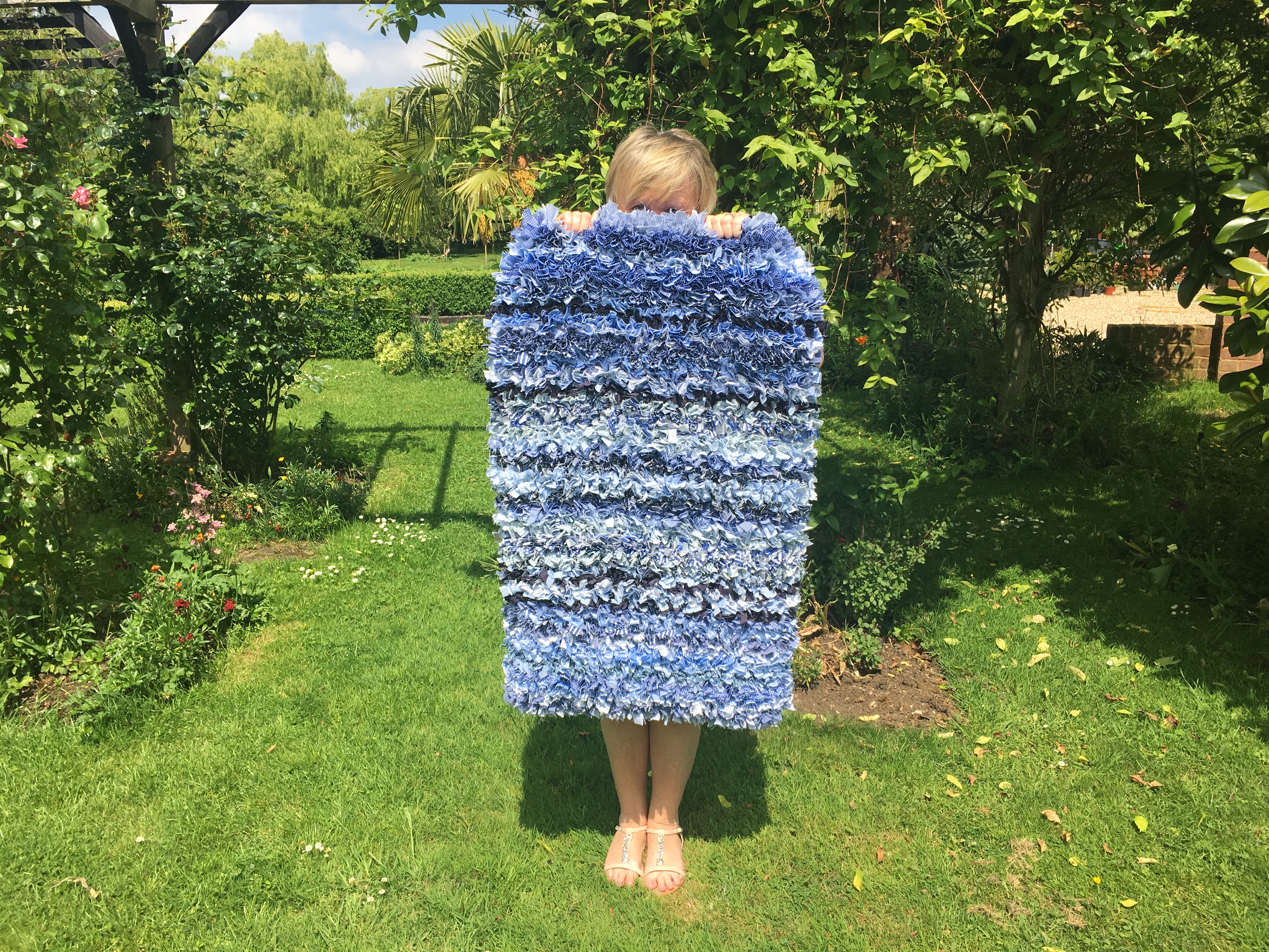 A woman holding a blue stripey shaggy rag rug in the garden