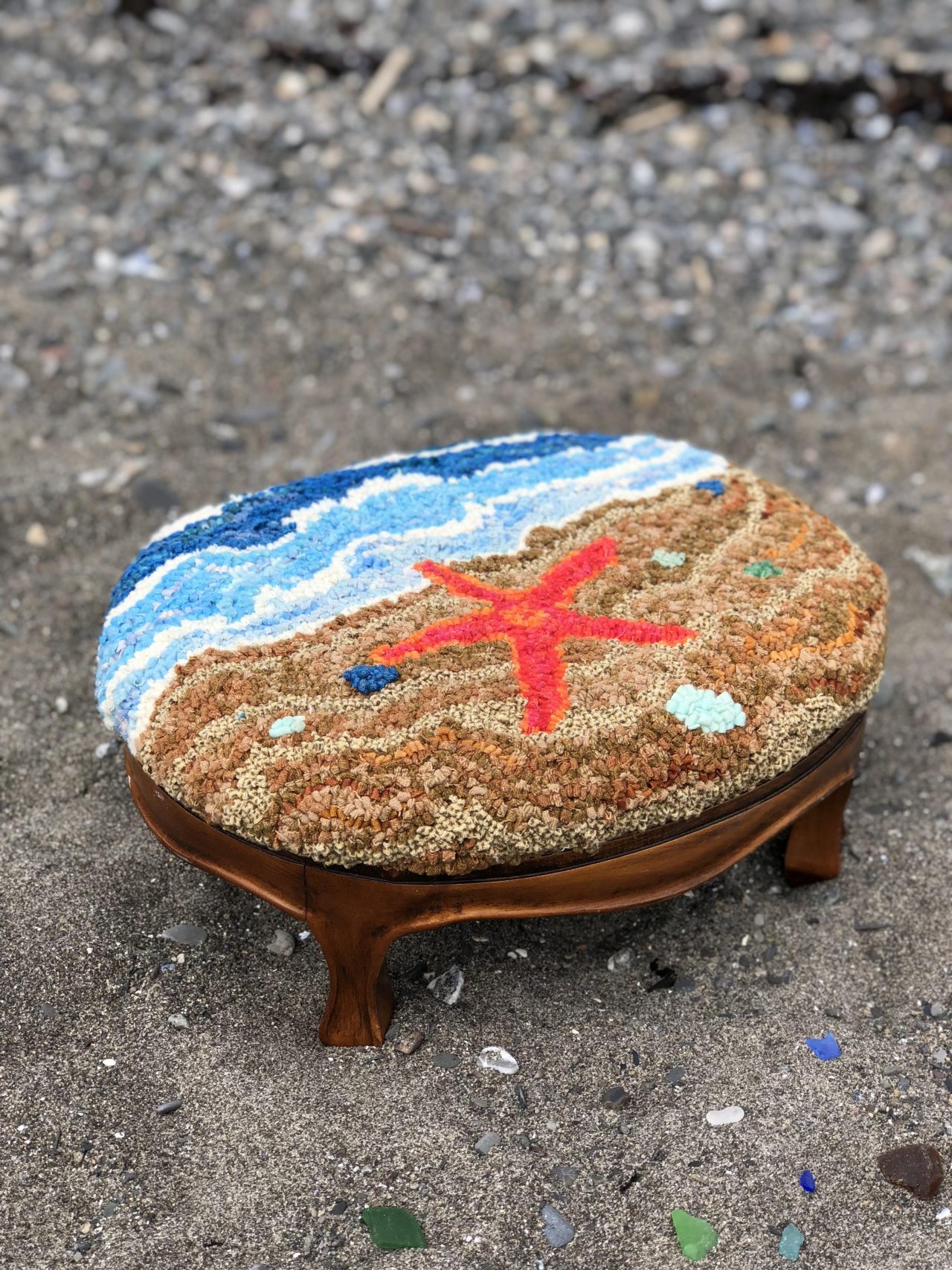 A wooden footstool with a hooked art image of a beach and red starfish by Yvonne Iten-Scott