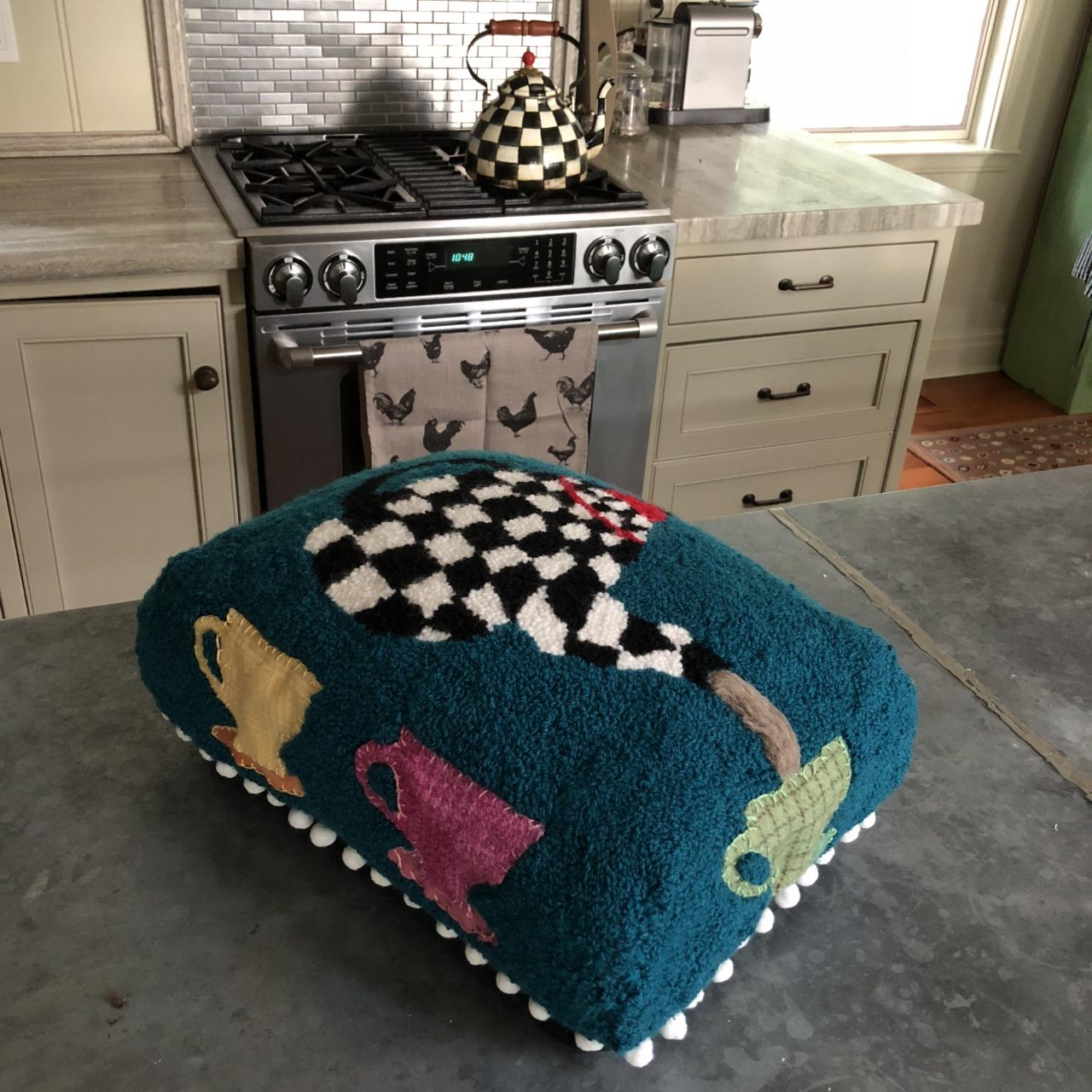 A textile art footstool with blue background, a black and white check teapot and coloured cups by Yvonne Iten-Scott