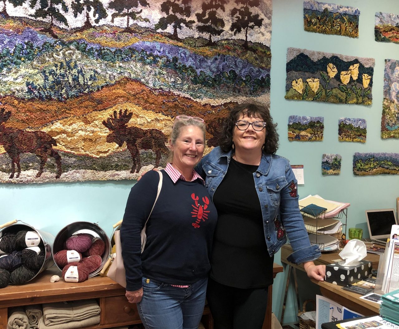 Yvonne Iten-Scott with Deanne Fitzpatrick standing in front of a wall of hooked art in Deanne's studio.
